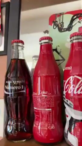 The video shows a shelf full of Coca-Cola bottles.  The bottles are decorated with different designs and labels, including city names like Napoli, Firenze, Milano, and Venezia, along with stylized illustrations of people.  Some bottles are transparent, showing the dark liquid inside, while others are painted a solid red color.  There are also Coca-Cola branded tins and other promotional items in the background.