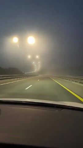 The video shows a first-person perspective from inside a vehicle driving on a highway at dusk or dawn. The road is well-lit by streetlights on tall poles that stretch into the distance. The sky is a gradient of deep blue and dark gray, with a faint haze or fog visible in the distance. Trees line the sides of the highway, and a guardrail is visible along the right side of the road. The dashboard of the car is visible at the bottom of the frame, with a black device, possibly a dashcam, mounted on it. The road markings include white lines separating lanes and a solid yellow line along the right edge. The vehicle appears to be moving at a moderate speed.