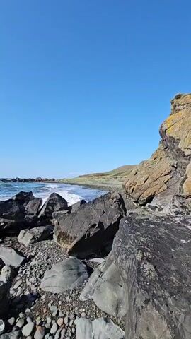 The video pans across a rocky shoreline under a clear blue sky. The ocean waves crash against the dark rocks, creating white foam. The shore is covered in a mix of large boulders and smaller pebbles. In the distance, a grassy hill slopes down to the sea. The sun is visible at times, reflecting off the water. There are no people or man-made objects visible in the video.