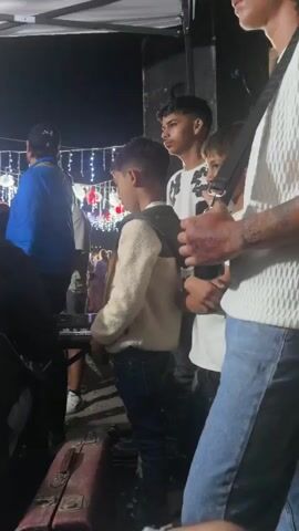 The video shows an outdoor gathering at night, likely a party or event. In the foreground, a young boy in a cream-colored jacket and jeans stands looking down, possibly at a keyboard or electronic device in front of him. Behind him, several other young people, mostly males, are visible. One person in a black and white patterned shirt is holding up a phone, appearing to record or take a photo. Further back, a group of people are watching a performance. On the right side of the screen, a person in a white shirt and jeans is partially visible. Towards the back, a drum set with multiple electronic pads is set up, and a drummer is actively playing. Decorative string lights are visible in the background, indicating a festive atmosphere. The scene is illuminated by artificial lighting, including spotlights directed at the performers. The overall impression is of a lively event with a musical performance taking place.
