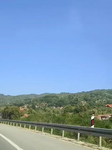 The video shows a view from a moving vehicle on a highway. The road is asphalt with white markings. A metal guardrail runs along the side of the road. Beyond the guardrail, there is lush green vegetation and trees. In the distance, rolling hills are covered in trees and dotted with houses. The sky is a clear, bright blue. A large blue sign with white text is visible on a gantry above the road. The sign appears to be a directional sign.
