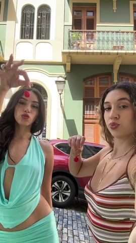 Two young women are standing on a cobblestone street in front of a building. The woman on the left is wearing a light teal two-piece outfit, and the woman on the right is wearing a striped tank top. Both women are holding cherries on strings. They playfully move the cherries towards their mouths, then make gestures with their hands and puckered lips. A red car is parked behind them. The building is painted in light green and beige colors, with ornate balconies and windows. The scene appears to be outdoors on a sunny day.