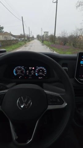 The video shows a first-person perspective from the driver's seat of a vehicle moving slowly down a narrow, wet road in a rural area. The sky is overcast, and there are puddles on the road. Houses and vegetation are visible on either side of the road, including power lines and purple flowers. The vehicle has a digital display showing speed and other information. The dashboard also has a large touchscreen display with a graphic of the vehicle. The gear stick and steering wheel are in view.