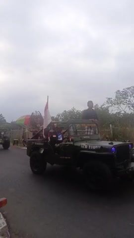 The video shows a parade of vintage jeeps on a paved road. The sky is overcast. In the foreground, a dark green jeep is prominently featured, with two people inside and one standing. The person in the driver's seat appears to be wearing a blue shirt. The person standing in the back is wearing a dark jacket and a cap. An Indonesian flag is mounted on a pole behind the driver. The jeep has the license plate "JGB 2109" visible. To the left of this jeep, another similar jeep is visible, and further behind it, a brightly colored umbrella or canopy is seen, along with some decorations and people. The road appears to be lined with vegetation on the right. A portion of a motorcycle is visible in the bottom left corner of the frame. The jeeps are moving forward in a procession.