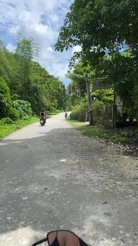 The video shows a person riding a motorcycle on a paved road surrounded by lush greenery. In the distance, another person is walking. A sign for "Southside Tourist Inn" is visible on the right side of the road. The sky is partly cloudy, and the overall atmosphere appears to be that of a tropical rural setting. The camera is mounted on the handlebars of the motorcycle, providing a first-person perspective.