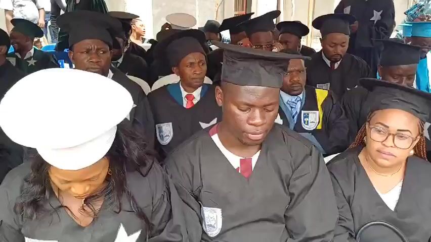 Several people, dressed in graduation gowns and mortarboards, sit in rows. Some are looking down, while others gaze forward.  A few wear glasses.  Some have ties. Some robes feature a patch.  Behind them is a makeshift structure, and some building windows. There is music in the background.