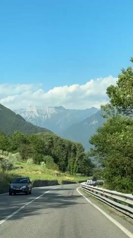 The video shows a scenic drive on a paved road that curves to the right, with mountains in the background. The sky is clear blue with some scattered clouds. On the left side of the road, there is a grassy slope with some trees and a stone wall. On the right side of the road, there is a metal guardrail and more trees. A white car is driving on the road ahead. The camera appears to be mounted on another vehicle, looking forward.