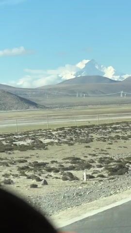 This video captures a vast, open landscape in Gyirong County, China, under a bright blue sky with scattered white clouds. In the distance, prominent snow-capped mountains dominate the horizon, their peaks sharply defined against the sky. Smaller, dark brown hills form a middle ground, partially obscuring the base of the larger mountains.

Across the midground, several power lines supported by transmission towers are visible, stretching horizontally across the scene. The foreground consists of arid, rocky terrain with patches of low-lying, scrubby vegetation. Several wooden poles, possibly part of a fence or another utility line, run parallel to what appears to be a road or track, only a sliver of which is visible in the bottom right corner, suggesting the video was shot from a moving vehicle. The overall impression is one of a remote, high-altitude environment with clear visibility on a sunny day. No people or visible text are present in the video.
