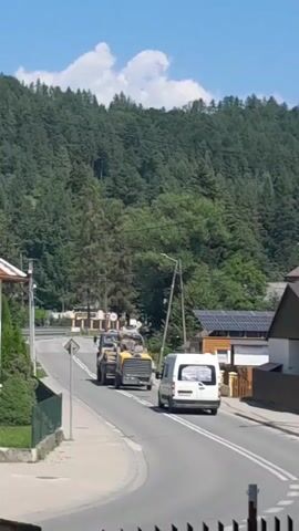 Two yellow construction vehicles, likely a tractor and a trailer, are driving down a paved road. A white van is following them. The road is bordered by houses and trees, with a dense forest covering a hillside in the background. The sky is blue with some white clouds.