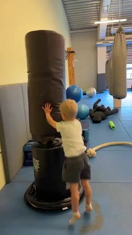 A young boy with blond hair, wearing a white t-shirt and gray shorts, stands on a blue mat in a gym. He is facing a large, black, free-standing punching bag and appears to be punching it. In the background, there are several other punching bags hanging from the ceiling, blue exercise balls, a black dummy, and some ropes and mats on the floor. The gym has a metal ceiling and fluorescent lighting. The boy is captured in motion, with his right arm extended towards the punching bag.