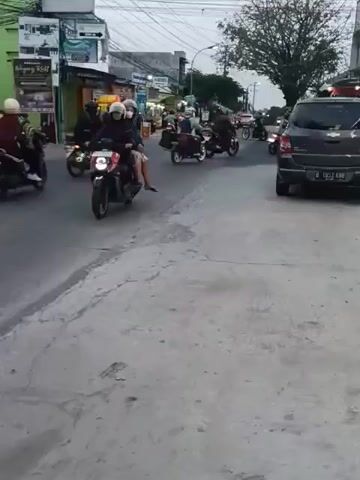 The video shows a street scene, likely in Southeast Asia given the presence of many motorcycles. In the foreground, a gray concrete walkway is visible, with a road running alongside it. Several motorcycles are seen moving along the road, some with passengers. On the left side of the road, there are buildings that appear to be shops or businesses, with signs in a foreign language. The sky is overcast, suggesting it may be early morning or late afternoon. The overall impression is of a busy urban or semi-urban environment.