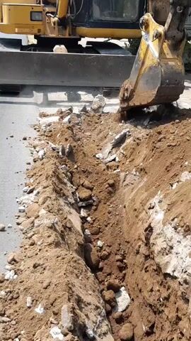 An excavator is digging a trench on a paved surface. The excavator is yellow and appears to be in operation. Dirt and debris are being removed from the trench, which is located to the right of the excavator. The trench is narrow and deep, with visible soil and rocks. The environment appears to be outdoors, possibly a street or construction site.