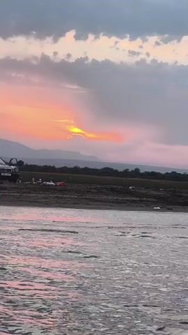 A truck and two SUVs are parked on a muddy shore near a body of water. Several people are standing or sitting near the vehicles. The sun is setting, casting a warm glow over the sky and mountains in the background. The water is calm with gentle ripples. The scene appears to be a casual gathering or picnic by the water.