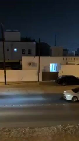At night, a video shows a street scene. Initially, a white car moves right to left on the road. Behind it is a light-colored pickup truck that is followed by another white sedan. The camera, originally focused on a low vantage point, slowly rises to reveal a perspective from the top of a building. Other cars can be seen in the distance heading towards a distant skyline of city lights. The video is shot with a steady hand, showing buildings and streetlights on either side of the road. The sky is a dark blue, characteristic of nighttime.