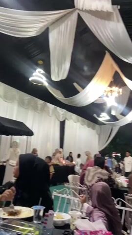 The video shows a dimly lit indoor event, possibly a banquet or reception, with guests seated at tables. The ceiling is decorated with flowing white fabric draped against a black background, and chandeliers are visible. In the foreground, a plate of food is prominently displayed, containing rice, a spicy red sauce with meat, a fried egg-like item, and crackers. Several stacks of clear plastic cups are arranged on the table. A person wearing a blue patterned shirt is seated at the table, looking towards the right side of the frame. Other guests are seen in the background, some wearing headscarves, and the overall atmosphere appears to be social and celebratory. The lighting suggests it is evening or indoors with artificial illumination.