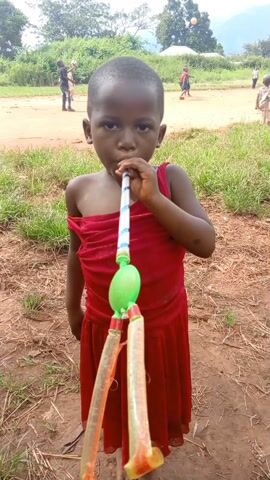 A young African child in a red dress stands in a dry, grassy area. The child blows into a homemade toy with a striped tube, a green bulbous part, and two clear tubes at the bottom. In the background, several people play on a dirt field. The child looks directly at the camera with a serious expression as they play with the toy. The surrounding environment includes green trees and a few distant hills.