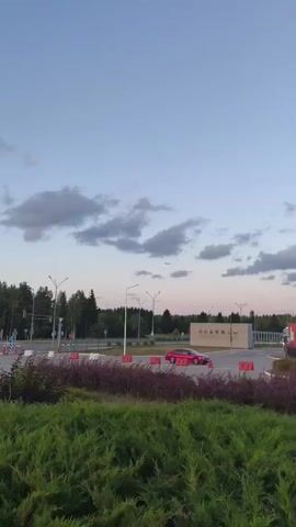 A video captures a pink car driving past a roadside in a suburban or rural area. Trees line the background with a few clouds in the sky. An airplane is visible in the distance. The shot appears to be taken from a slightly elevated position, looking down on the scene.