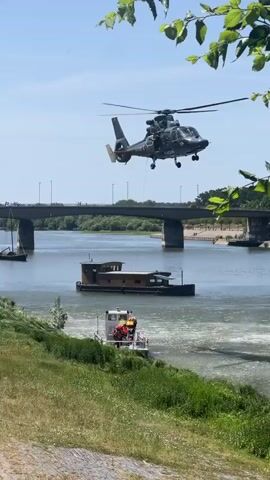 A black helicopter hovers over a wide river. Below the helicopter, a rescue operation is underway. A person in a yellow harness is being lifted from a darker boat by the helicopter. On a smaller white boat in the foreground, two individuals in red vests are observing the operation and appear to be assisting with a rope. A large, dark barge or house-like boat is stationary in the river between the rescue boat and the far bank. A bridge spans the river in the background, with trees lining the banks. The sky is clear and blue, indicating a sunny day.