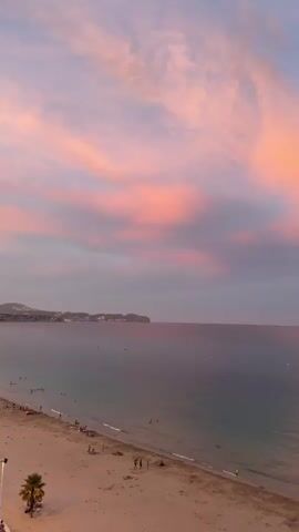 The video captures a serene beach scene at sunset. The sky is ablaze with vibrant pink and orange hues, painted across a canvas of soft purple and blue clouds. The ocean, a deep, tranquil blue, gently laps at the shore. On the sandy beach, a few individuals are scattered, some enjoying the water, others relaxing on the sand. A lone palm tree stands on the left side of the frame, adding a tropical touch to the idyllic setting. The overall atmosphere is one of peace and natural beauty.