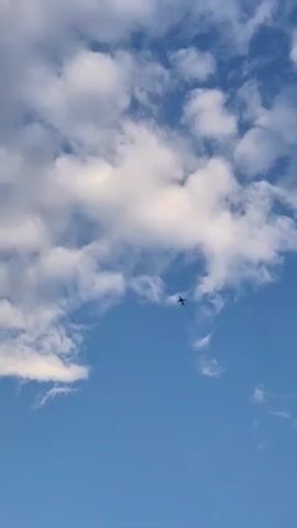 The video captures a clear blue sky dotted with fluffy white clouds. A small airplane is visible in the distance, soaring through the sky. The camera is positioned to look upwards, focusing on the vast expanse of the sky and the distant aircraft. There are no people or ground-level objects visible in the frame. The lighting suggests it is daytime, likely with sunlight illuminating the clouds.