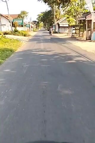 The video captures a first-person perspective, likely from a motorcycle, traveling down a paved road in a rural or semi-rural area. The road is bordered by vegetation and buildings. Shadows of trees are cast across the road, indicating it is daytime, likely morning or late afternoon due to the angle of the sun. On the left side of the road, there is a green sign with text in Indonesian. Further down the road, a person is riding a motorcycle in the same direction as the camera. On the right side of the road, there are small buildings, some with open fronts that appear to be shops or homes. The road surface shows some wear and tear, with tire marks visible in some sections. The overall impression is a mundane journey through a village or small town.