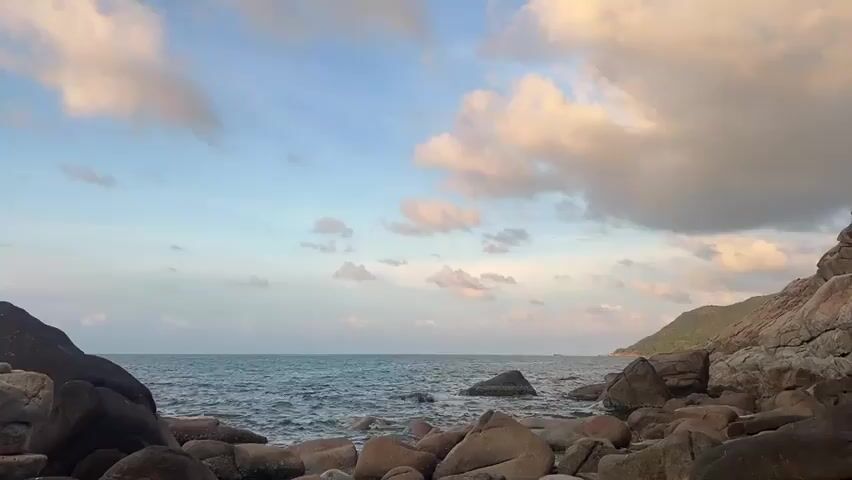 The video showcases a rocky coastline on a partly cloudy day. The foreground is filled with various sized rocks, some wet from the water. A small cairn of stacked rocks sits to the left. The ocean stretches out to the horizon, with a slight wave action visible. In the distance, a green, hilly landmass extends out into the sea. The sky is a blend of light blue and white clouds, some tinged with a hint of pink. The overall scene is calm and natural.