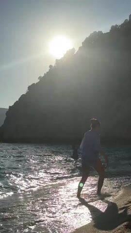 A vertical video captures a tranquil beach scene at what appears to be sunset or late afternoon. The bright sun hangs low over a dark, rocky cliff that dominates the background, casting a strong backlight and brilliant reflections on the gently rippling ocean waves.

In the foreground, a person with reddish hair, wearing an oversized light blue shirt over a dark swimsuit bottom, walks from the sandy shore into the shallow water. They turn slightly, seemingly adjusting their hair or clothing, before continuing to face the sea. Further out in the water, another person is partially visible as a dark silhouette, already immersed.

The water is clear, and the waves are small, creating sparkling patterns as they catch the sunlight. The overall atmosphere is peaceful and serene, with the strong sun creating dramatic contrasts between light and shadow. The location context is Kaş, Turkey. No visible text is present in the video.