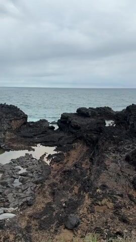 The camera pans from the horizon over the ocean to reveal a rocky shoreline. The rocks are predominantly dark with some areas of brown and beige. There is a small pool of water nestled among the rocks. In the distance, a massive cliff covered with green vegetation rises from the sea. Clouds obscure the top of the cliff. Waves are crashing against the base of the cliff. In the last part of the video, a person with red hair comes into view on the right-hand side of the screen. The weather appears to be overcast and possibly damp.