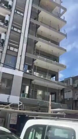 The video shows a street scene in an urban environment. In the first shot, a tall building under construction is visible, with multiple balconies. Scaffolding is present around the base of the building. There is a white van parked in front. The next shot pans up to show a blue sky with scattered clouds and several other buildings. A construction worker in a blue helmet and light green vest is visible. The final shot shows a row of parked cars and a large bush with yellow flowers along a sidewalk. Other people are visible walking in the distance.