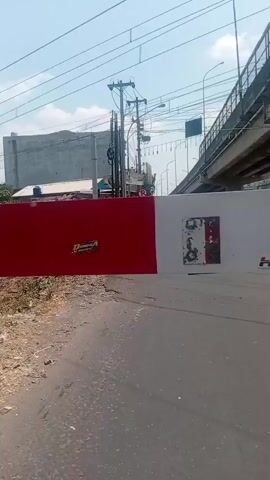 A white and red railroad crossing gate is in the foreground, partially obscuring a train. The train is white with red and grey stripes and has the letters "KAI" on the front. The train moves from left to right across the screen. In the background, there are buildings, utility poles, and overhead wires. The sky is clear and blue.