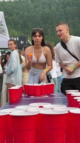 A woman in a white bikini top and a light blue wrap skirt throws a small orange ball towards a table set up with red plastic cups. She is positioned behind the cups and is looking at them intently. A man in a white t-shirt stands to her right, looking at the camera with a smile. The table is covered in rows of red cups, suggesting a game of beer pong. In the background, other people are visible, some standing and others seated. The setting appears to be outdoors, with trees and a mountain visible in the distance. There is a white tent structure on the left side of the frame.