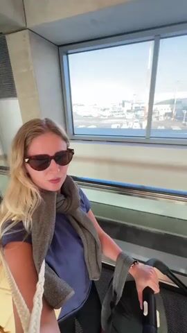 A woman with blonde hair and sunglasses is seen on an escalator at an airport. She is wearing a blue t-shirt, a gray scarf, and is carrying a yellow bag over her shoulder. She is also holding onto the escalator handrail and appears to be looking towards the camera. In the background, through large windows, an airport tarmac is visible with airplanes, buildings, and control towers under a clear blue sky. The overall impression is that of a traveler moving through an airport, possibly heading for a flight, with a sense of anticipation or preparedness. The time of day appears to be daytime, with bright sunlight indicating fair weather. There is no visible text in the media.
