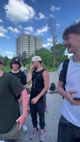 A group of young men are gathered in a parking lot with a tall building in the background and trees on the right. The sky is blue with white clouds. One of the men, shirtless and wearing black pants, is holding a megaphone and speaking to the group. The other men are dressed casually in t-shirts, hoodies, and baseball caps, and some are holding cell phones. Several scooters are visible on the ground. The lighting suggests it is daytime.