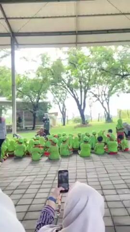 The video shows a group of children sitting on a paved surface under a covered area. They are mostly wearing green shirts with red pants. Several adults, primarily women wearing hijabs, are standing around them. One adult appears to be instructing or organizing the children. Some people are holding and using mobile phones, potentially recording or taking pictures of the scene. The background features trees and open green space. The setting looks like an outdoor gathering or field trip.