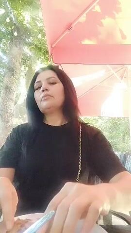 A woman with long black hair and dark eyes, wearing a black t-shirt, sits at an outdoor table under a pink umbrella. She is holding a slice of pizza with both hands and bringing it to her mouth to take a bite. Her head is tilted slightly as she eats. There are lights strung in the background, and foliage is visible behind her. The video is filmed from a close-up perspective, focusing on the woman and her food.