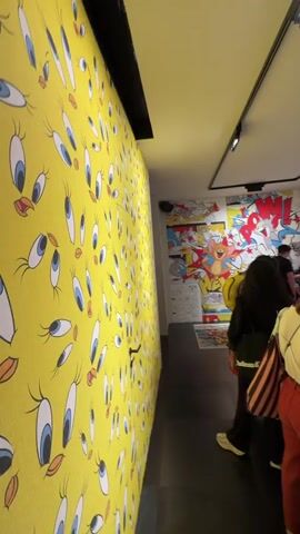 This video captures an indoor space, likely a store or exhibition, with vibrant Looney Tunes themed decor. A prominent wall on the left features a repeating pattern of Tweety Bird's eyes against a yellow background. The opposite wall is adorned with colorful murals depicting various Looney Tunes characters such as Bugs Bunny, Road Runner, Wiley Coyote, Sylvester the Cat, and Daffy Duck, in different scenes, including a "POW!" graphic.

Several people, mostly adults, are present. They are actively engaged in browsing and examining items, which appear to be wallpaper samples, design books, or catalogues, displayed on tables in the center of the room. Some individuals are standing, while others are seated on low chairs or benches. One person is seen filming with a phone. The floor features a large circular rug with a target-like design incorporating Looney Tunes characters. The lighting is bright, provided by track lights on the ceiling. The overall atmosphere suggests a casual browsing experience in a creatively themed environment.