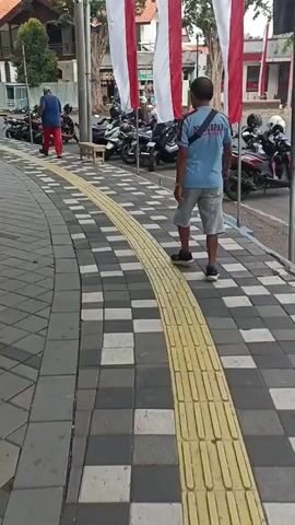 A man in a light blue t-shirt and grey shorts walks away from the camera down a checkered sidewalk. A yellow tactile paving strip runs down the center of the sidewalk. Behind him to the left, several motorcycles are parked. Further left, a man in a blue shirt and red pants walks away from the camera. To the far left, several people are walking along the sidewalk, and a black car is parked on the street. To the right of the man in the blue t-shirt, more motorcycles are parked. In the background, buildings with orange tile roofs and trees are visible. Three large red and white flags hang vertically on poles to the right of the man.