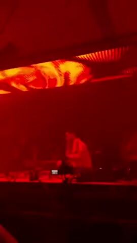 A DJ in a white Adidas track suit stands behind a lighted sound board on a stage and raises his right hand. Behind the DJ are several people silhouetted against a bright light. The stage has metal supports.
