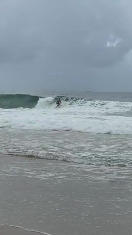 A person skimboards on a breaking wave at a beach. The individual, wearing dark shorts, is positioned on a skimboard, riding the white foam of a wave that breaks from left to right. In the foreground, shallow water ripples on the sandy shore. The ocean extends into the background, where several small boats are visible on the distant horizon beneath a heavily overcast sky.