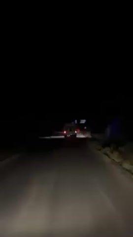 Here is a description of the video:

The video shows a vehicle driving on a dark road at night. The headlights of the car illuminate the road ahead, and the taillights of the vehicle are visible in the distance. The surroundings are dark and blurry. The video seems to be taken from inside another moving vehicle. The overall atmosphere is one of isolation and darkness.