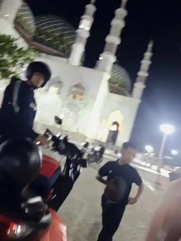 In this video, three young people are gathered outside a mosque at night. The mosque, with its white walls, multiple minarets, and green domes, is illuminated by artificial lights. 

On the left, a young person wearing a black hoodie and helmet is seated on a motorcycle. In the center, a boy in a black t-shirt and pants holds a helmet. To the right, a girl in a light pink hijab and a blue skirt stands, holding her hands up near her face. 

The ground is paved with cobblestones, and several other motorcycles are parked nearby. Streetlights cast a warm glow on the scene. The overall mood appears casual and social, with the young people seemingly interacting with each other. The time of day is clearly night due to the darkness and the artificial lighting. The presence of the mosque suggests a location of religious significance. No visible text is present in the video.