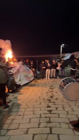 A group of people, including a man beating a large drum, gather at night in a stone-paved courtyard, watching a fire with fireworks erupting from it.  There are several other people standing around the fire holding up smoking pyrotechnics.  A white banner with writing on it sits in front of the fire. The night sky is dark, with a single street lamp visible in the background.  Many people are holding phones up, presumably recording the scene.  The atmosphere appears festive.