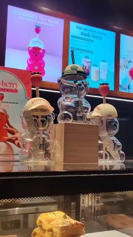 The video pans across a display of three clear glass bear-shaped Starbucks tumblers. The tumblers are designed to resemble teddy bears and have straws with strawberry-shaped toppers. Two tumblers are placed on either side of a central tumbler, which is elevated on a wooden block. Each tumbler has a small Starbucks logo. Behind the tumblers, there are illuminated signs with promotional images of Starbucks drinks and text. One sign features a pink bear tumbler with a strawberry topper, and the text "Your Drink's New Bestie." Another sign displays various Starbucks drinks with the text "Your everyday choices, made better." The bottom of the frame shows a display case with pastries. The lighting suggests an indoor setting, likely a Starbucks store.