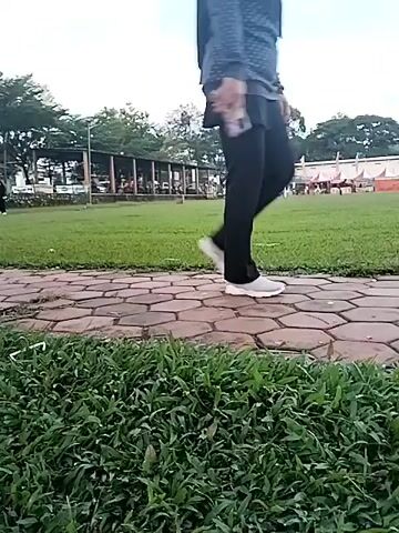 This video captures an outdoor scene, likely a public park or recreational area, in Matesih, Indonesia. The setting features a large, well-maintained grassy field bordered by a paved path made of hexagonal bricks in the foreground. In the background, there are numerous trees, some providing shade, and several simple buildings with brown roofs, possibly small cafes or kiosks. A soccer goal is faintly visible towards the right side of the field in the initial frames.

The time of day appears to be daytime, possibly late afternoon, given the bright yet somewhat diffused light and the partly cloudy sky.

At the 0:01 mark, a person wearing a gray and black long-sleeved shirt, black patterned pants, white shoes, and a hijab is seen walking on the paved path. They are holding a phone or a small recording device up in their right hand, seemingly taking a selfie or recording their walk.

Throughout the video, several other people are visible in the background, walking around the field and near the buildings, suggesting a communal gathering or leisure activity. The camera pans slowly, offering different perspectives of the area. No specific event is clearly identifiable, but the overall activity is casual strolling and enjoying the outdoor space. There is no clearly readable text visible in the video.