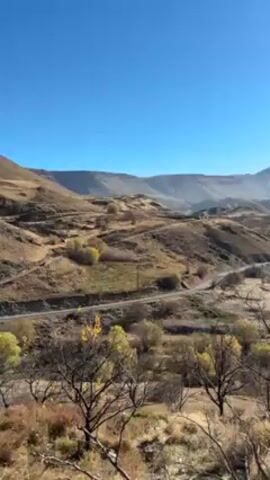 The camera pans across a sunlit, hilly landscape with a road winding through it. The hills are mostly brown and dry, with some sparse vegetation. The sky is clear blue. As the camera pans, a valley comes into view, with a stream and a few trees. Smoke is visible in the distance.