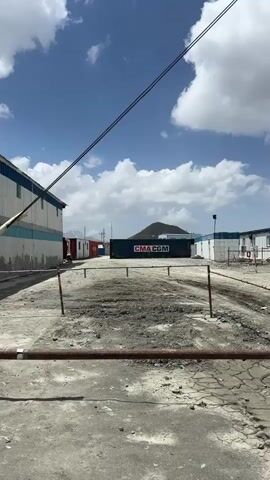 The video shows a wide outdoor area under a cloudy blue sky. In the foreground, a cracked dirt or gravel surface with tire tracks is visible, bordered by a red and white striped barrier on posts. A brown metal pipe or bar runs horizontally across the lower portion of the frame.

To the left, a long, light-colored building with a blue roof and windows runs along the edge of the frame. Further back, another building with a blue roof and white walls is visible.

In the center, a large dark blue shipping container with white lettering that reads "CMA CGM" is prominent. Behind the container, a dark, hilly landscape with power lines and structures on its peak can be seen.

To the right of the shipping container, another building with a blue roof and white walls is visible, with some smaller structures and a white tire next to it. In the background, to the right, a white tanker truck with lettering on its side is parked on a road.

The scene appears to be an industrial or logistical yard, possibly a port or distribution center, with evidence of construction or maintenance in the foreground. The lighting suggests it is daytime. There are no people visible in the video.