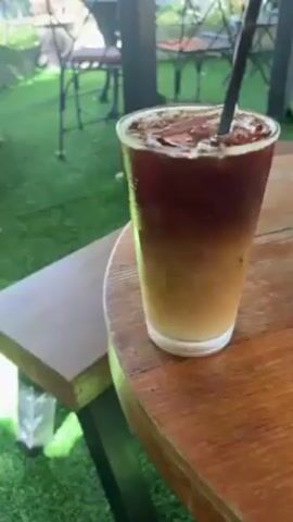 The video shows a tall glass of iced coffee on a round wooden table outdoors. The coffee is layered, with a dark brown liquid at the top and a lighter tan liquid at the bottom. Ice cubes fill the top portion of the glass, and a black straw is inserted into the drink. A hand is visible, stirring the coffee with the straw. In the background, there is green artificial turf, a wooden bench, and black metal chairs and tables.