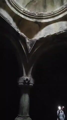 The video shows the interior of a stone structure with arches and a partial dome. The dome has layers of circular decoration, with a hole in the top allowing a small amount of light in. The stone appears aged and cracked, with some green moss growing on it. Below, arches are supported by a pillar with a decorative design at its peak. In the dark background, two figures can be seen. One is holding a light source, while the other is only partially visible. The lighting is dim, with the dome illuminated by the small opening above.