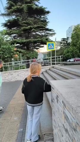 A man and a young girl are walking down a sidewalk near a pedestrian crossing. The man is pulling two large suitcases behind him. The girl is walking ahead of him, looking to her right. Another girl is standing on the sidewalk near a lamppost, looking out at the water. There is a body of water in the background, with a pier and some buildings visible. The sky is clear and blue.