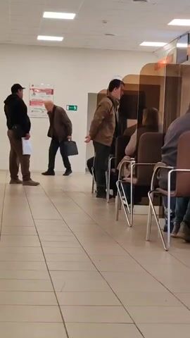 Here is a description of the video: 

The video shows the interior of what appears to be a waiting room in an office or government building. The room has a tiled floor and a suspended ceiling with fluorescent lights. The walls are white.  On the left, two men are standing and talking. One man is wearing a black jacket, brown pants, and a black baseball cap, holding some papers. The other man has on a brown jacket, dark pants, and is carrying a black bag. To the right, a row of chairs is lined up with people sitting in them. Clear plastic dividers separate the chairs, presumably to provide a barrier between people.  A poster is visible on the wall to the left.  An exit sign is also visible on the wall above the seated people.  A man in the middle of the line of chairs is talking to someone in front of him.  The overall impression is of a bureaucratic setting where people are waiting for assistance or services.